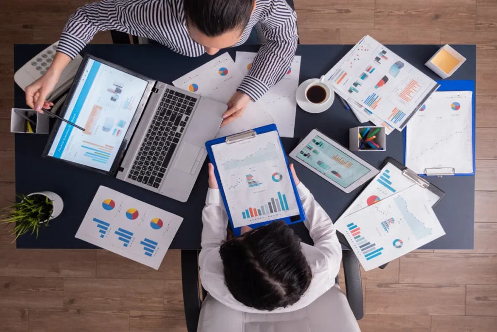 Visão de cima de duas pessoas em uma mesa, analisando relatórios e gráficos de endomarketing em um laptop, tablet e pranchetas.
