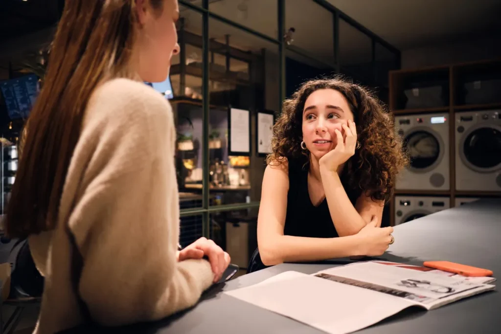 Duas jovens sentadas à mesa, uma olhando com uma expressão distante enquanto a outra parece falar com um semblante preocupado e a mão no rosto, sugerindo uma possível falta de empatia na interação.