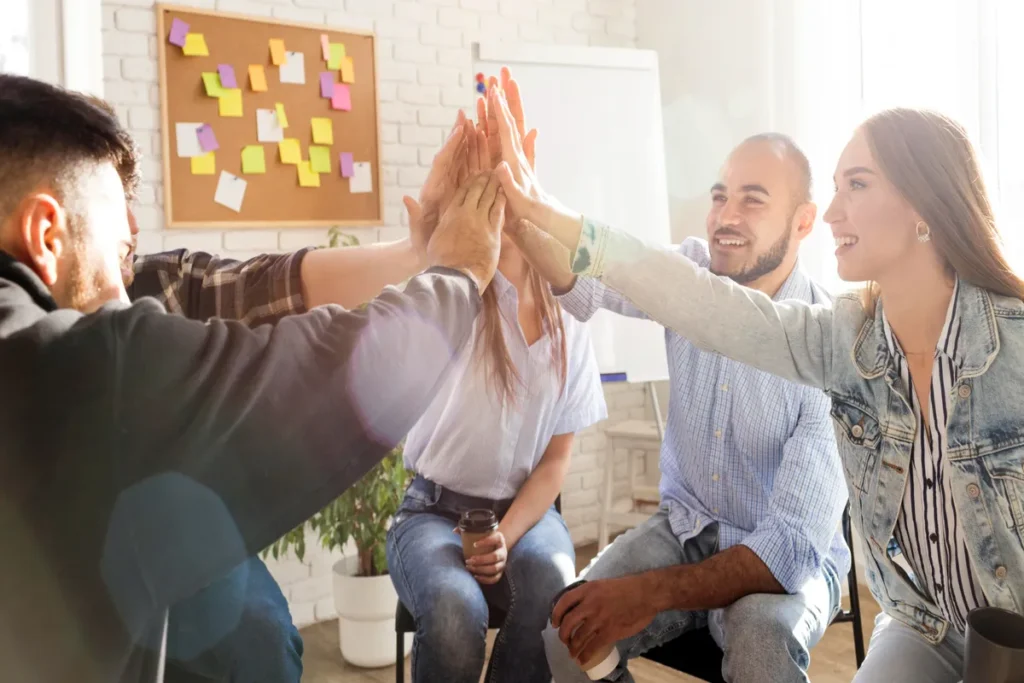 Cinco colegas de trabalho sorrindo e celebrando um sucesso com um high-five em grupo, com as mãos unidas no centro de um círculo, em um escritório, ilustrando o tema 'incentivo' e união de equipe.