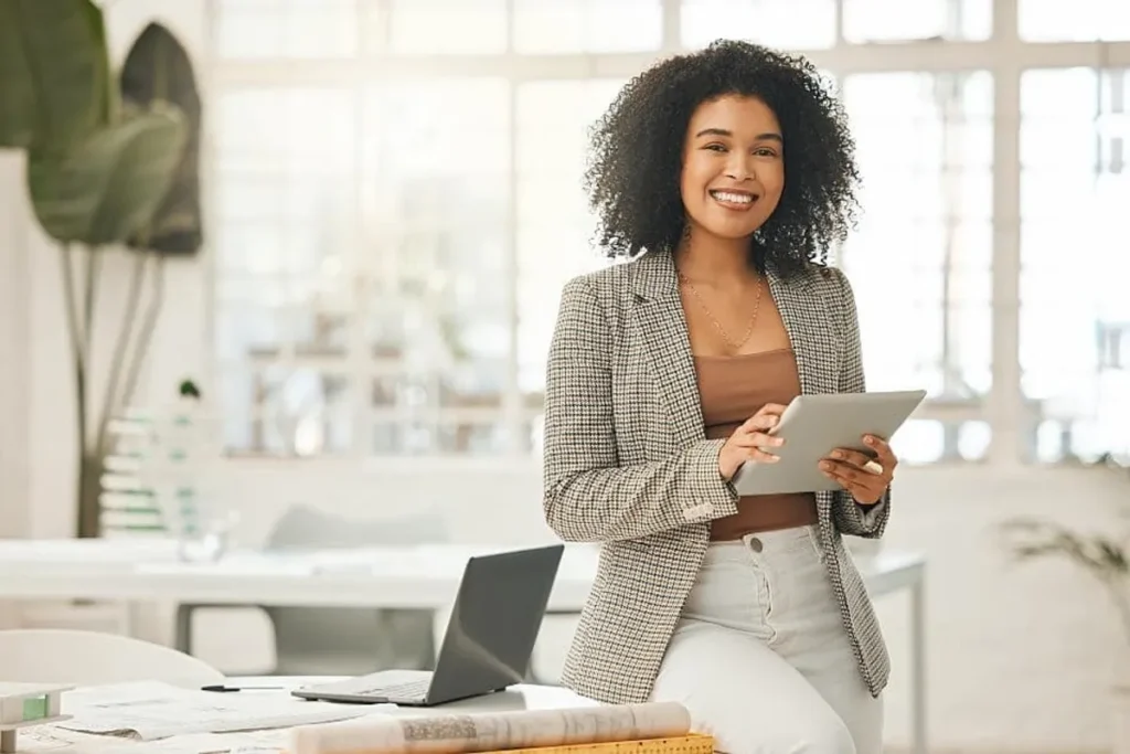 Uma mulher jovem, negra, de cabelo cacheado, sorri para a câmera enquanto segura um tablet, vestindo um blazer xadrez e calças claras em um escritório moderno e iluminado.