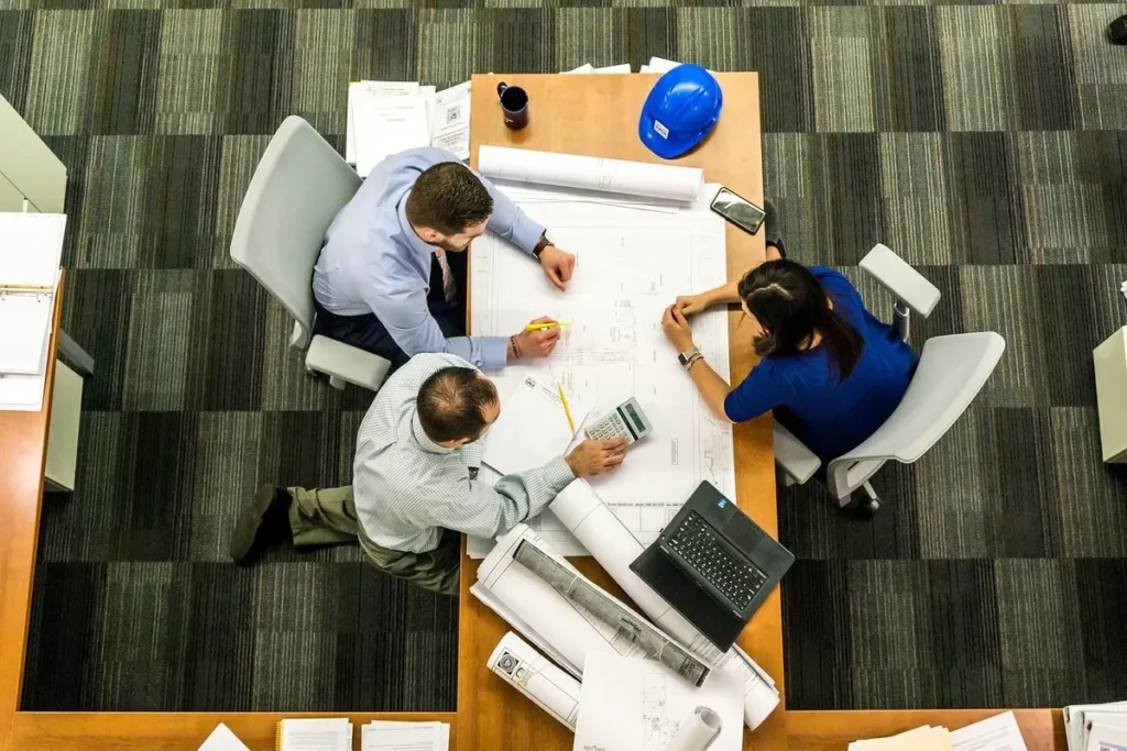 Três profissionais em reunião de engenharia, analisando plantas arquitetônicas, discutindo e identificando lacunas em treinamento de equipe.