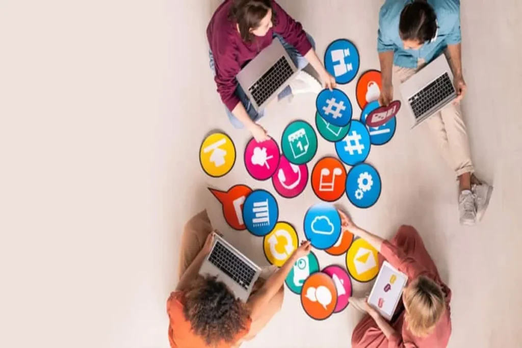 Três pessoas sentadas no chão, usando laptops e tablets, cercadas por grandes ícones coloridos de redes sociais e marketing digital. A imagem representa o planejamento de estratégias de marketing de relacionamento.