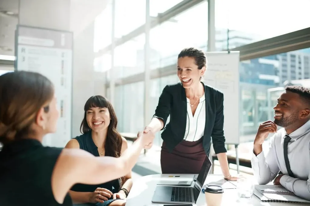 Uma mulher de blazer preto apertando a mão de outra pessoa em uma mesa de reunião, com outros dois colegas sorrindo ao lado. A cena representa a construção de parcerias e o marketing de relacionamento em um ambiente corporativo.
