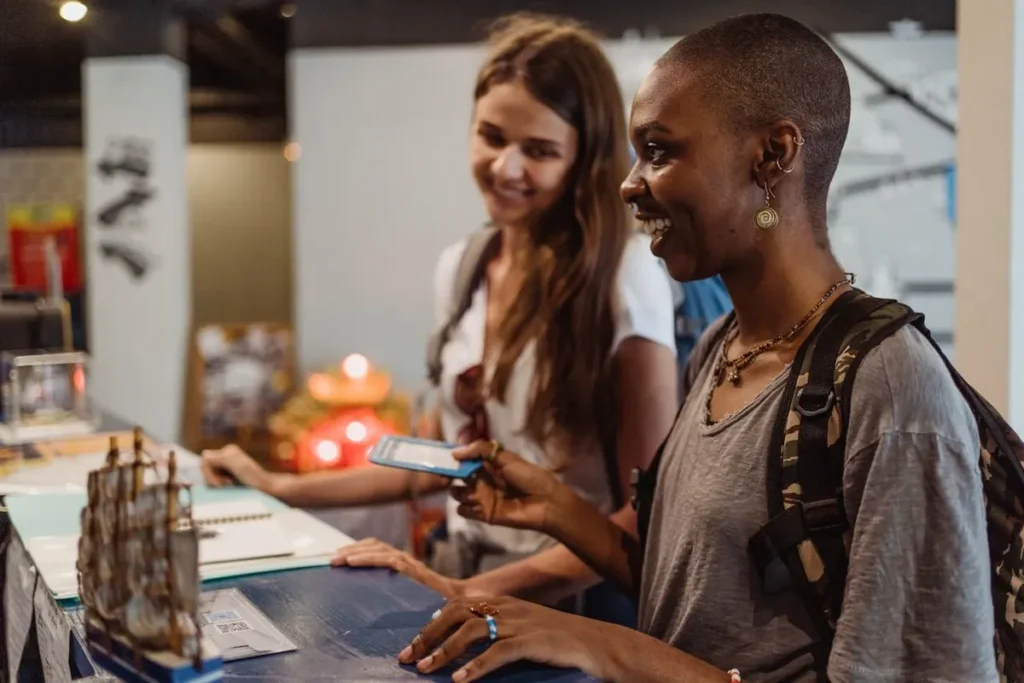 Duas mulheres sorridentes em um balcão de atendimento padronizado, uma delas com cabelo raspado e mochila segurando um cartão azul, a outra ao fundo com cabelos longos e mochila.