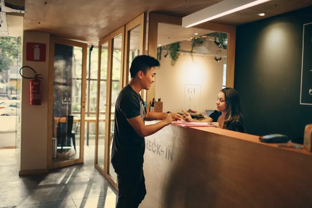 Atendimento padronizado em uma recepção ou balcão de check-in, com um homem de camiseta preta interagindo com uma mulher que está atrás do balcão de madeira, que possui a palavra "CHECK-IN" gravada.