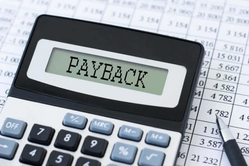 Uma calculadora em cima de uma folha de cálculo financeira, com a palavra "PAYBACK" a ser exibida no ecrã. A imagem ilustra o conceito de retorno de investimento.