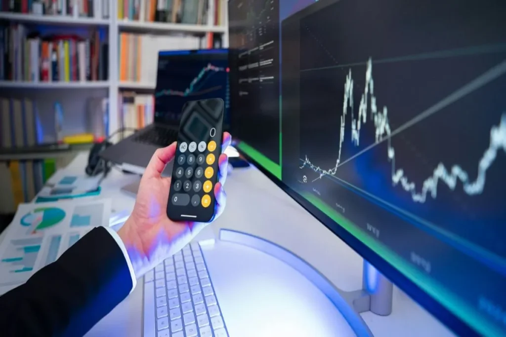 Uma mão segura um smartphone com uma calculadora, enquanto um monitor ao fundo mostra gráficos financeiros de investimento, representando o conceito de payback.
