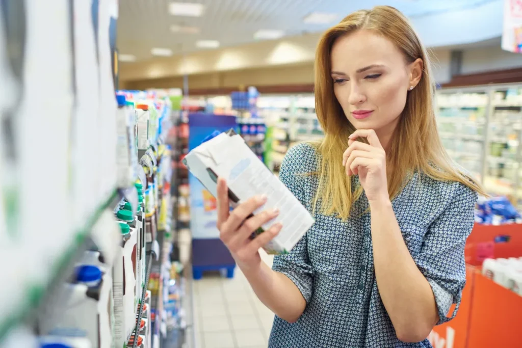 Mulher loira em supermercado examinando a embalagem de um produto, atenta à sua precificação ou informações nutricionais.