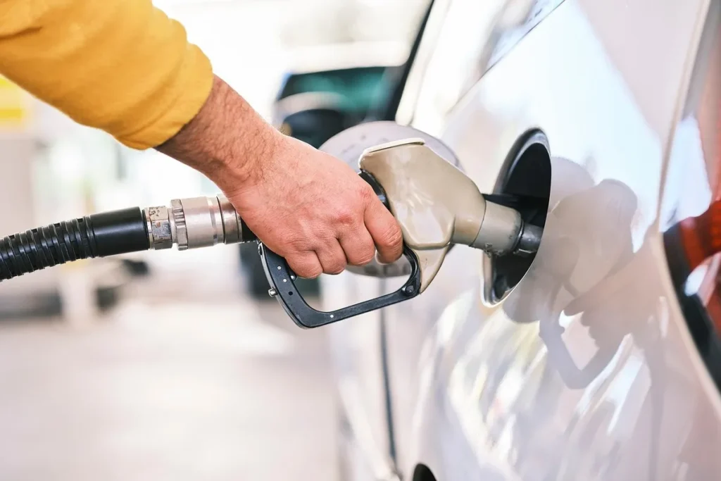 Mão de uma pessoa segurando um bico de abastecimento inserido no tanque de um carro branco, ilustrando o serviço diário nas redes de postos.
