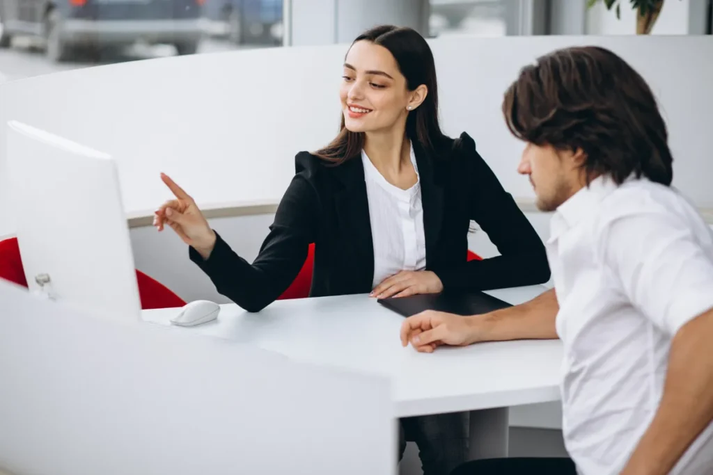 Uma mulher de blazer preto e camisa branca, sentada ao lado de um homem, aponta para a tela de um computador, sorrindo. A cena demonstra "relacionamento com o cliente", consultoria e atendimento personalizado em um ambiente de escritório moderno.