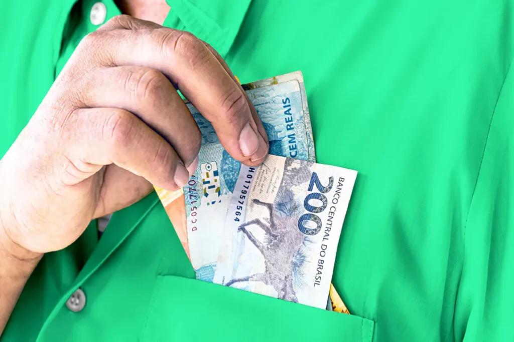 A mão de uma pessoa colocando notas de R200eR100 no bolso de uma camisa verde, representando o conceito de renda extra.