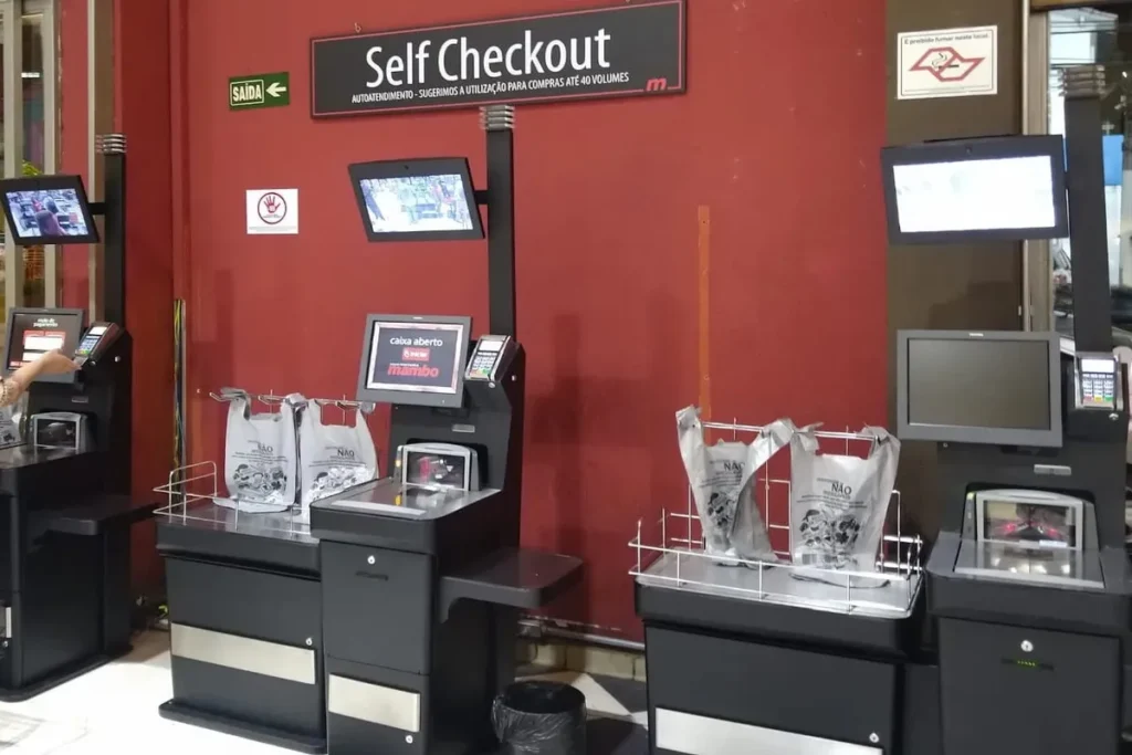 Fileira de terminais de self-checkout em um supermercado com sacolas e uma pessoa usando o serviço, ilustrando o autoatendimento.