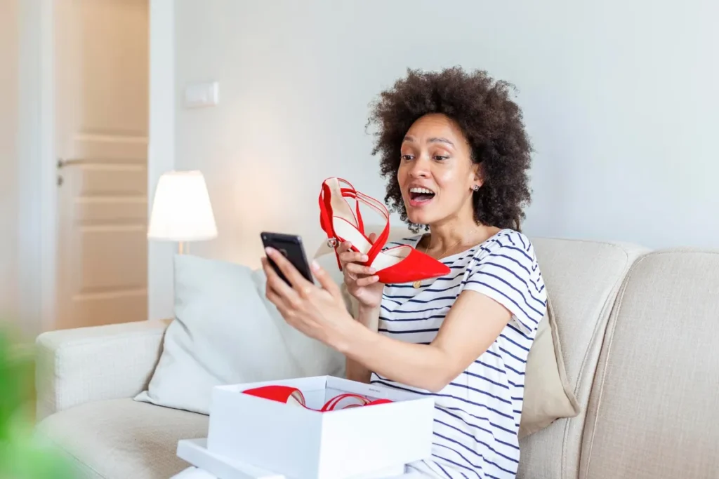 Mulher negra com cabelo afro, sorrindo e surpresa, tirando um par de sapatos de salto alto vermelhos de uma caixa, representando o momento de "unboxing".