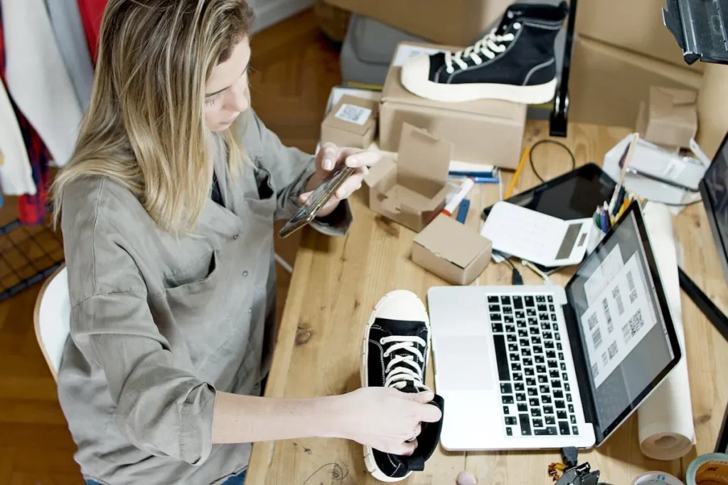 Mulher fotografando um tênis com o celular em uma mesa de trabalho, com um laptop e caixas de entrega, representando o processo de varejo online.