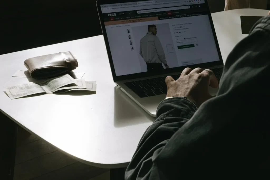 Pessoa comprando um casaco no site ASOS em um laptop, com carteira e dinheiro na mesa, ilustrando o processo de varejo online.