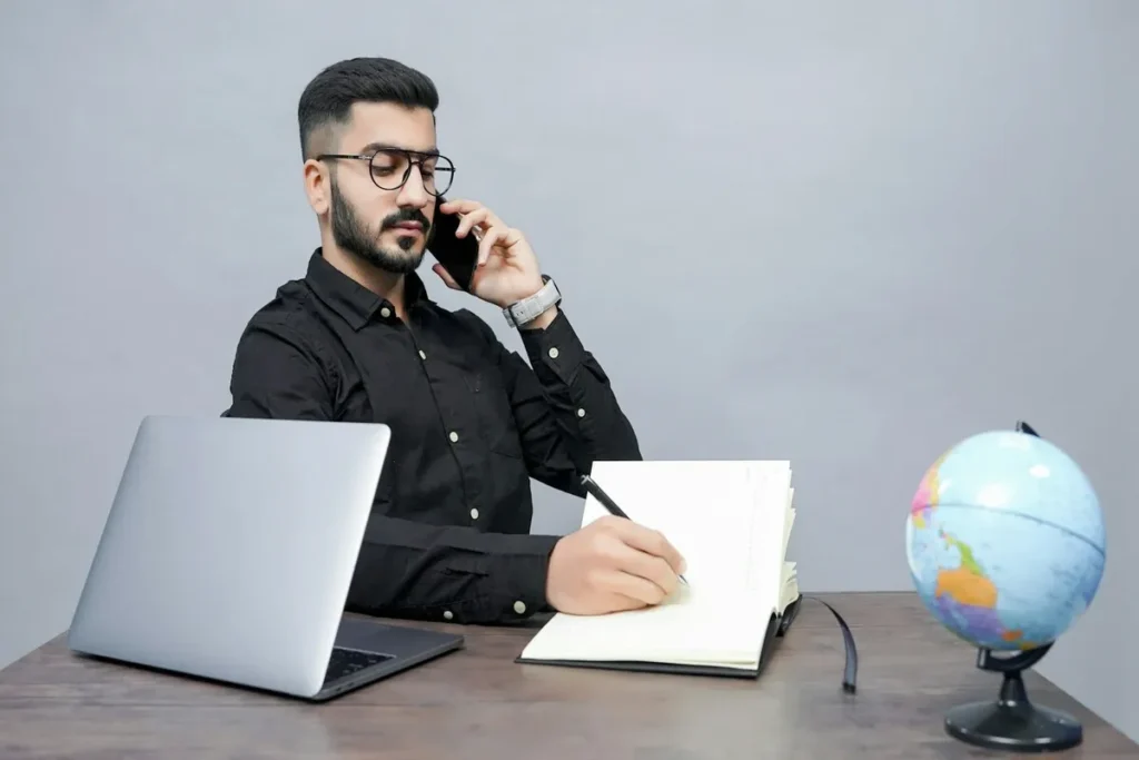 Homem de óculos, barba e camisa preta em call, anotando informações em caderno, com laptop e globo terrestre sobre a mesa, representando a venda consultiva e o trabalho remoto.