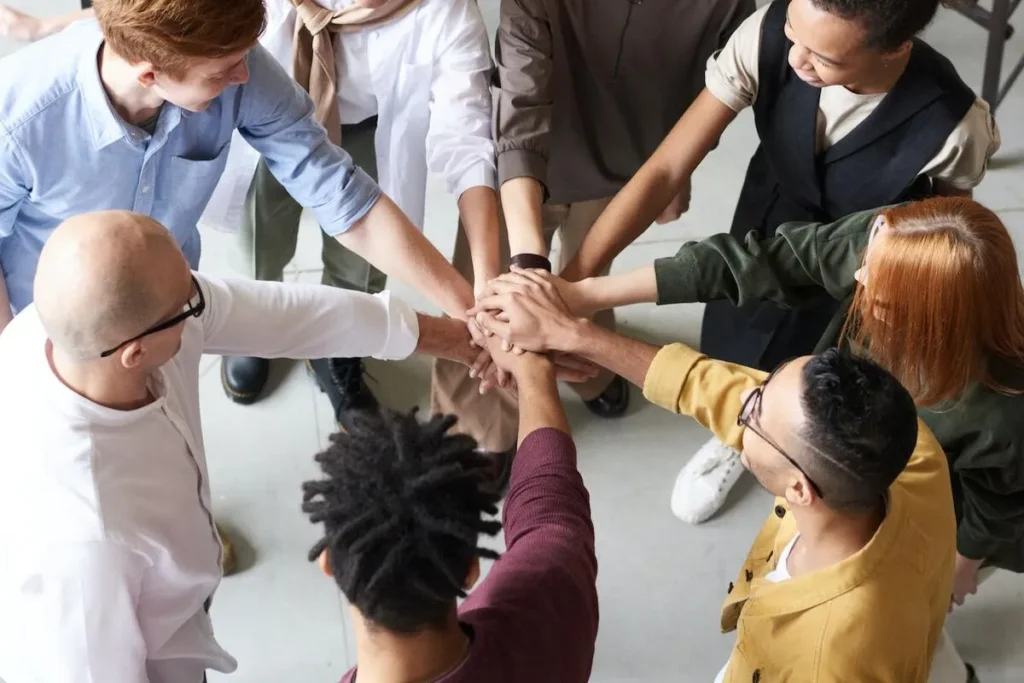 Um grupo diversificado de profissionais, incluindo homens e mulheres de várias etnias, empilha as mãos em um gesto de união e colaboração, ilustrando o conceito de endomarketing e trabalho em equipe em um ambiente corporativo.