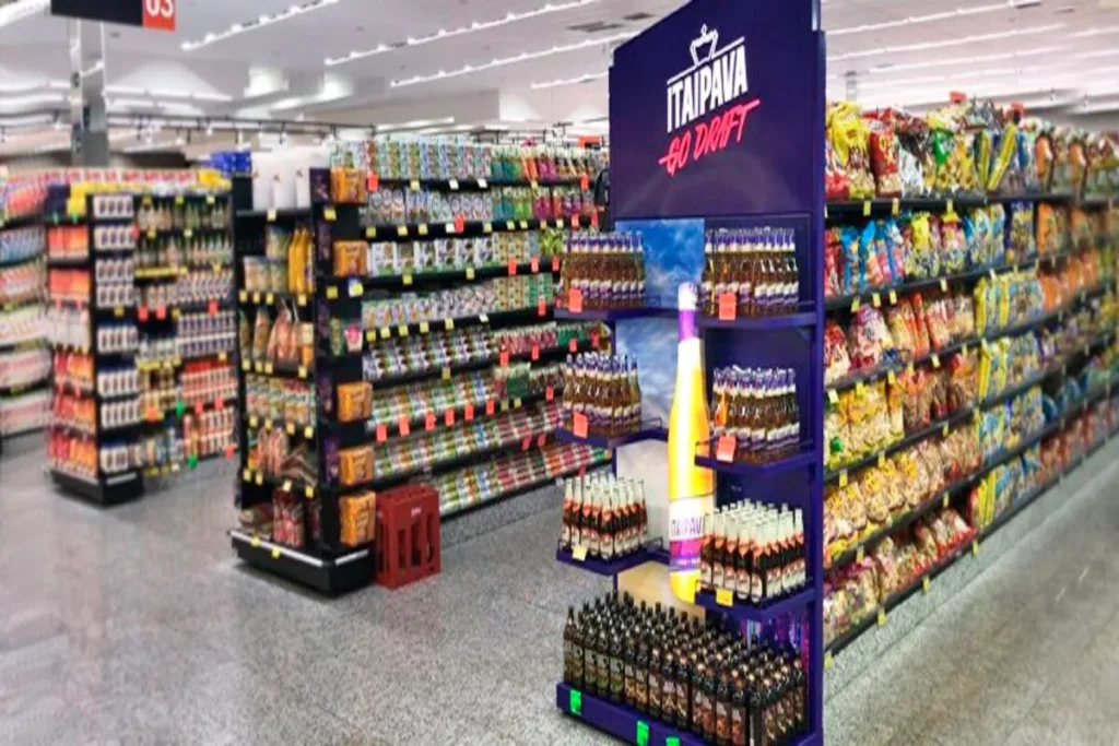 Imagem de um corredor de supermercado, com prateleiras cheias de produtos, e um MPDV (Material de Ponto de Venda) em destaque com a marca Itaipava. A imagem ilustra a aplicação de estratégias de MPDV para chamar a atenção do consumidor.