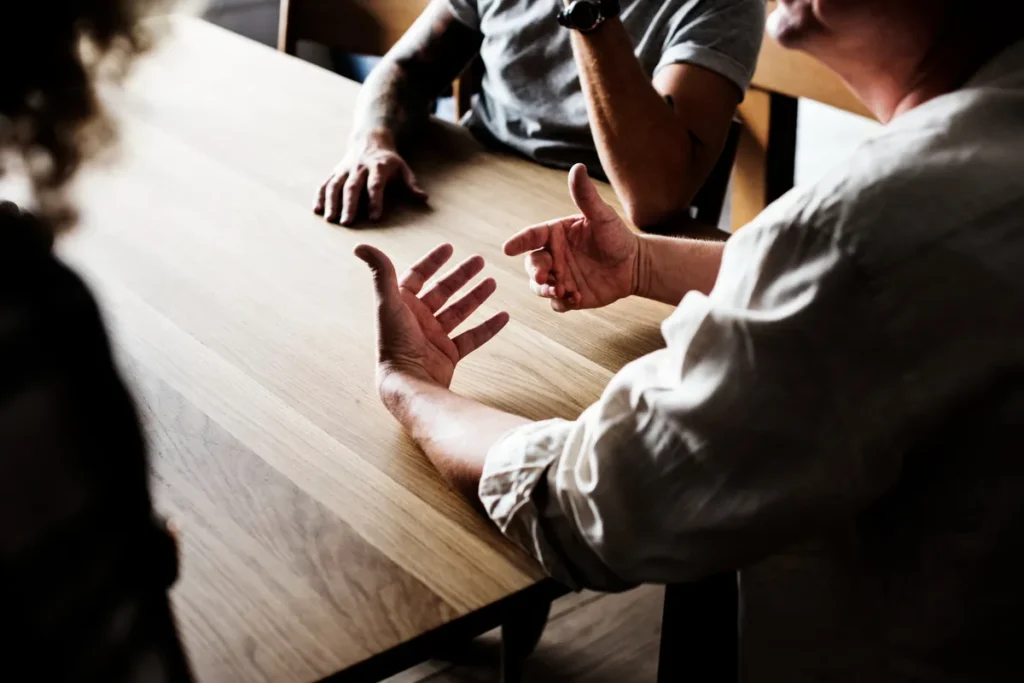 Pessoas sentadas em uma mesa, gesticulando com as mãos durante uma discussão de negócios ou argumentação.