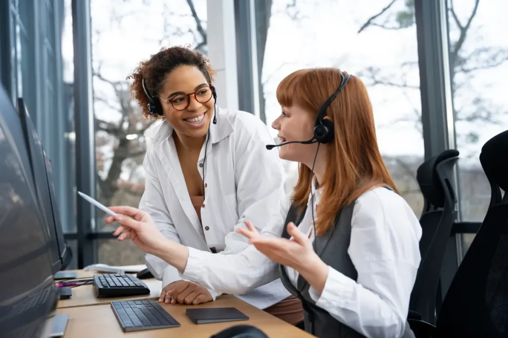 Duas mulheres sorridentes em um escritório, uma auxiliando a outra em um computador, com a palavra-chave "atendimento humanizado".