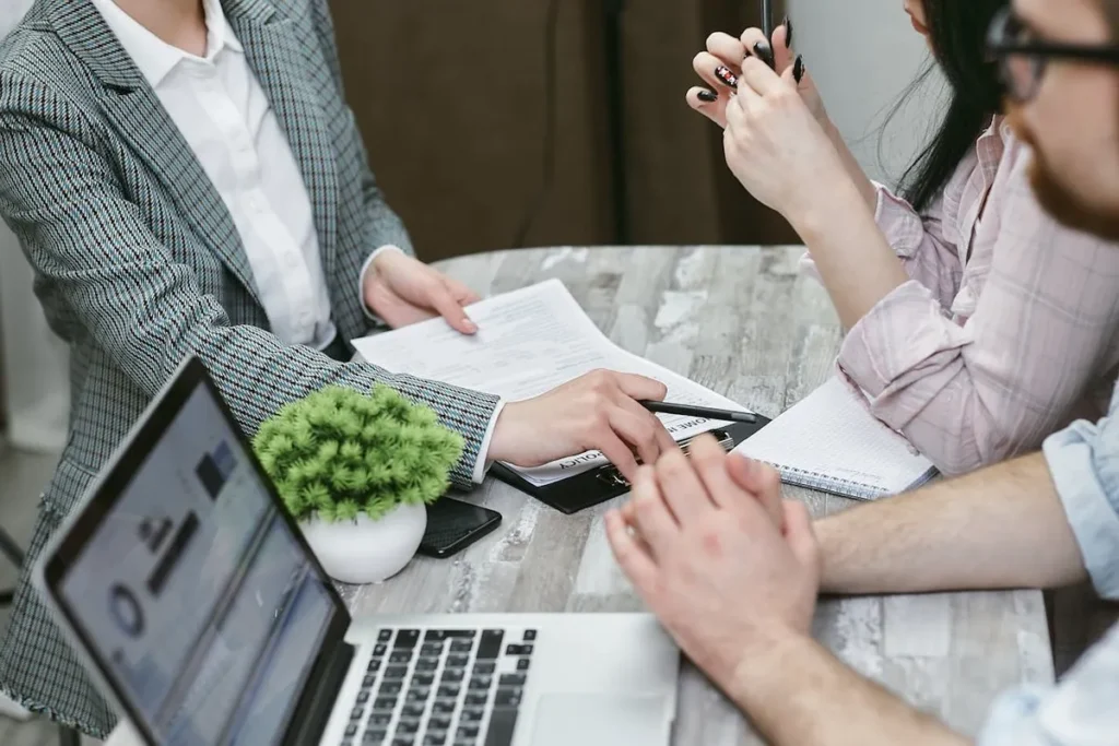 Três pessoas em uma mesa, discutindo documentos e dados em um notebook, em um encontro de capacitações profissionais ou de consultoria.