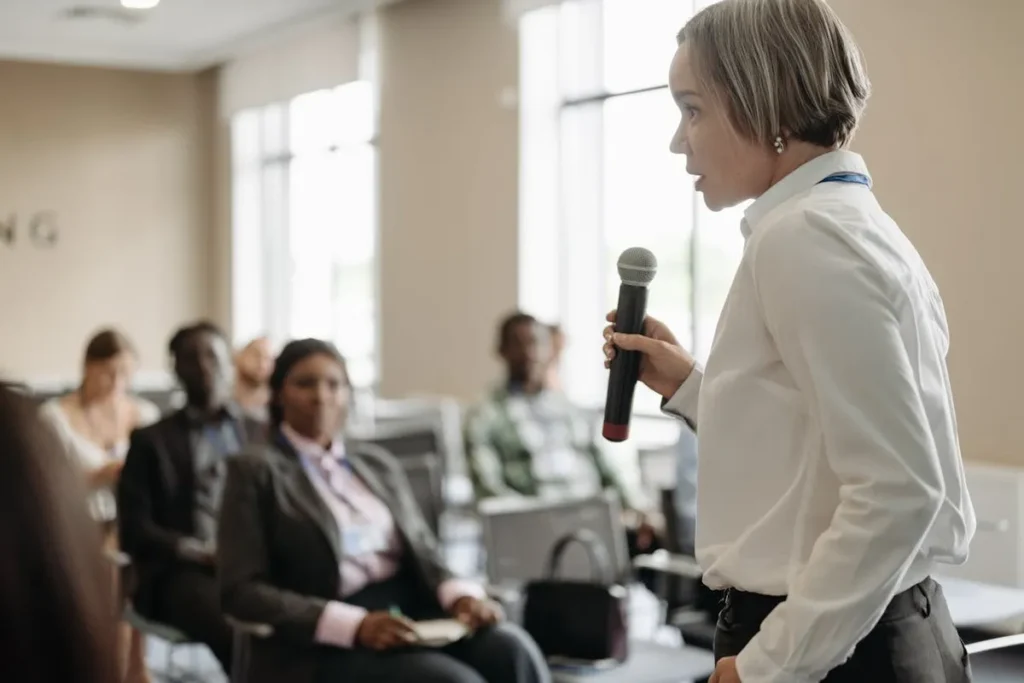 Mulher segurando um microfone e falando para uma plateia em um evento de capacitações profissionais.