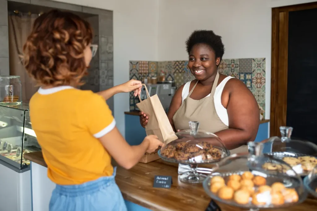 Mulher dona de uma padaria sorri enquanto entrega um pedido para uma cliente, lidando bem com a concorrência.