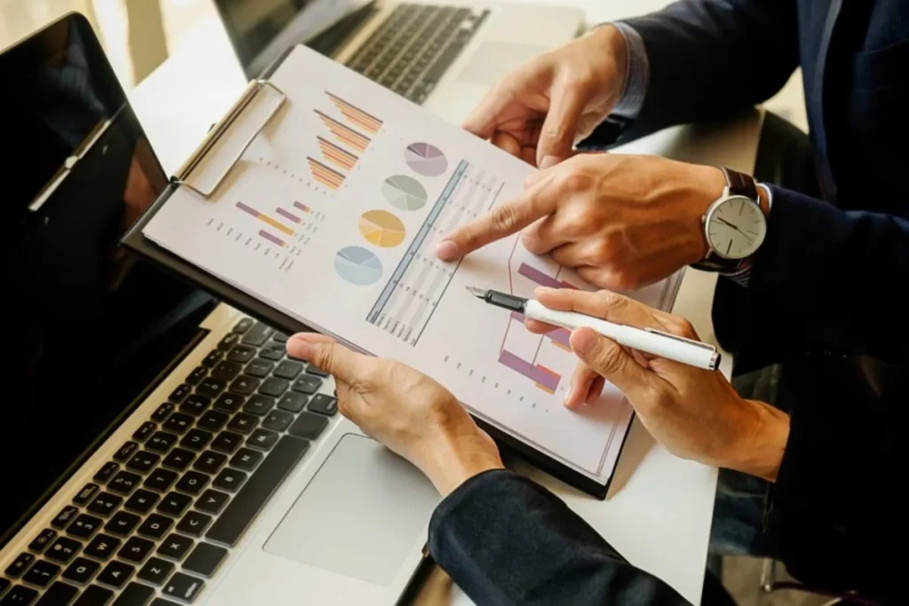 Duas pessoas em um escritório, em um close-up de suas mãos, analisando gráficos e relatórios financeiros em uma prancheta, simbolizando a busca por conformidade e resultados.