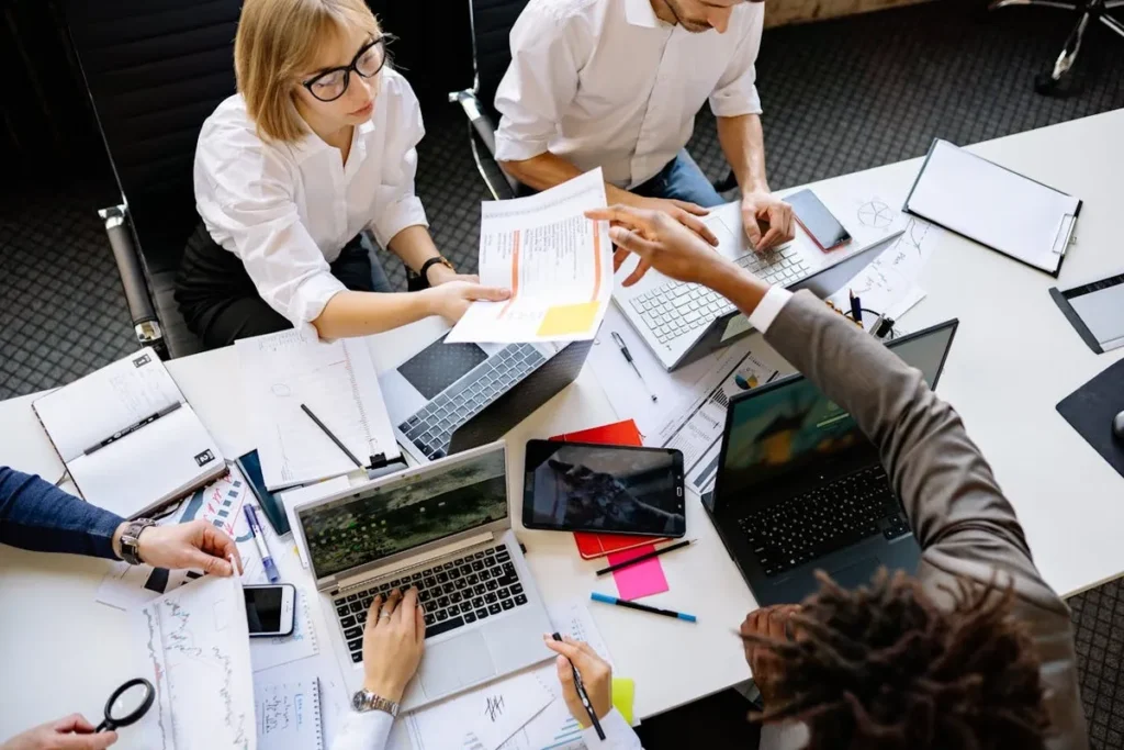 Vista aérea de uma mesa de reunião com diversas pessoas trabalhando em laptops e analisando documentos, com uma atmosfera de colaboração intensa.