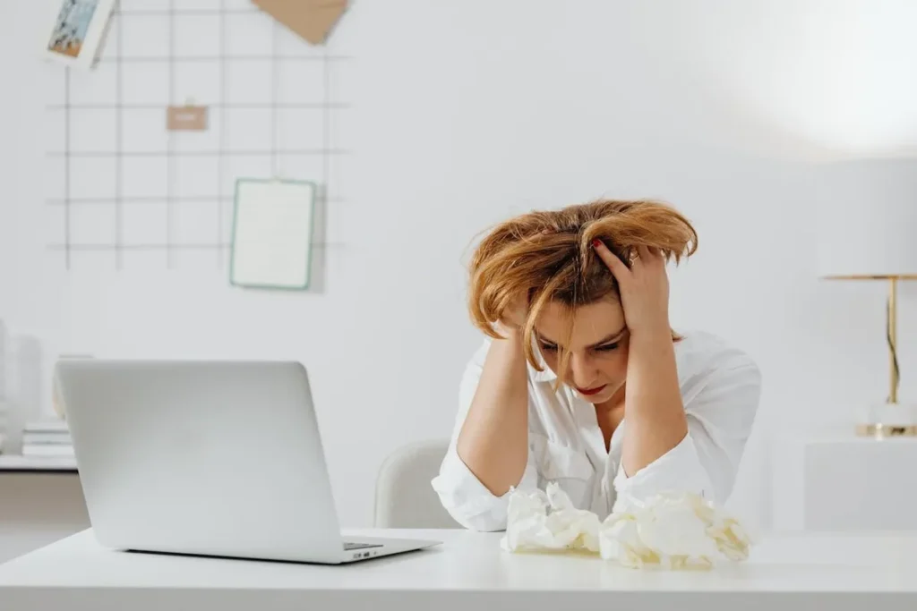 Mulher loira com expressão de cansaço e desânimo, segurando a cabeça com as mãos em uma mesa de escritório com um notebook e papéis amassados, simbolizando a desmotivação profissional.
