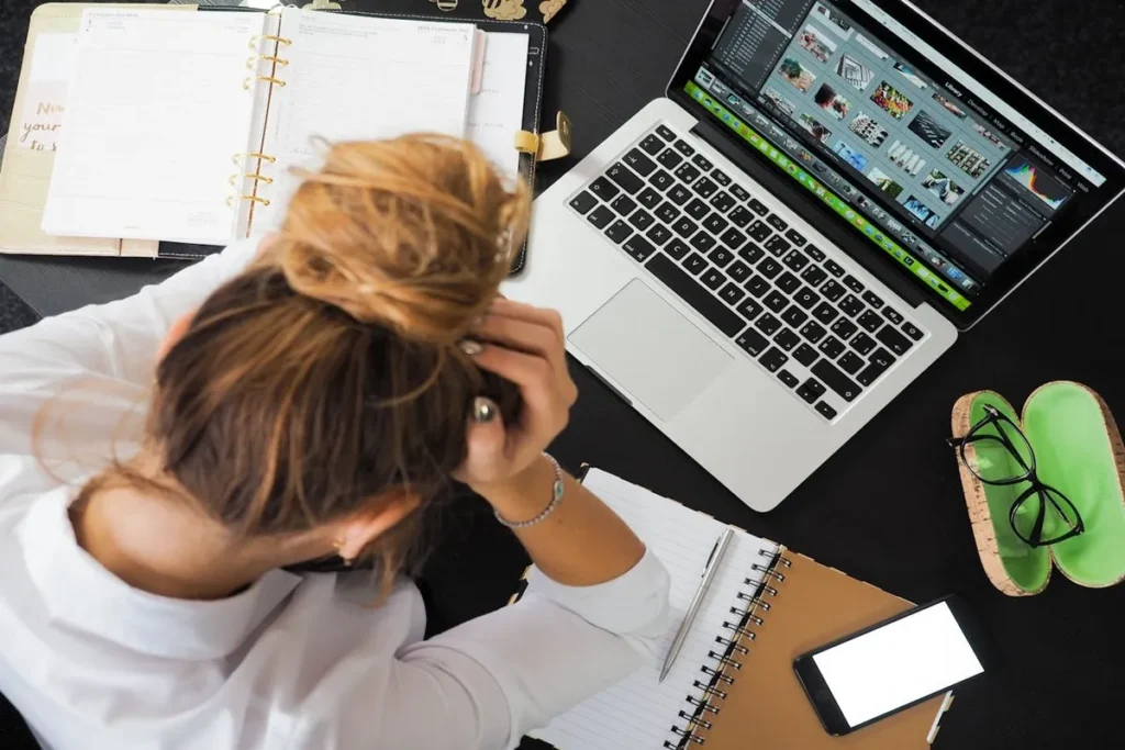 Vista de cima de uma mulher em uma mesa de escritório com a cabeça entre as mãos, ao lado de um laptop, óculos e papéis, expressando exaustão e desmotivação.