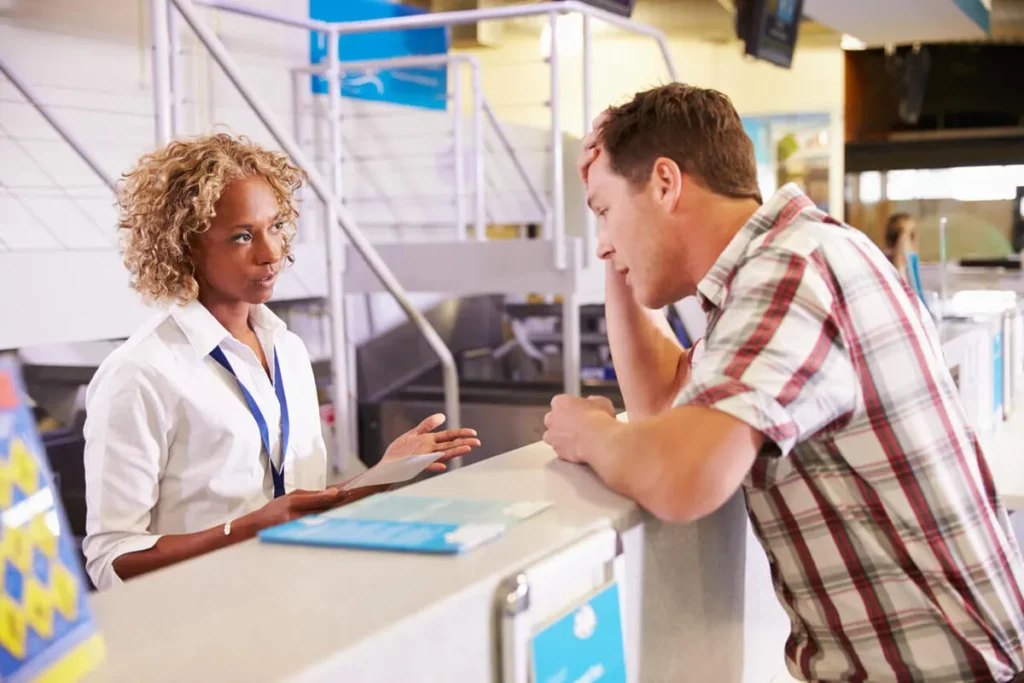 Funcionária de atendimento em um balcão de aeroporto conversando com um passageiro frustrado, que tem a mão na cabeça, ilustrando uma falha no serviço ou um problema de viagem.