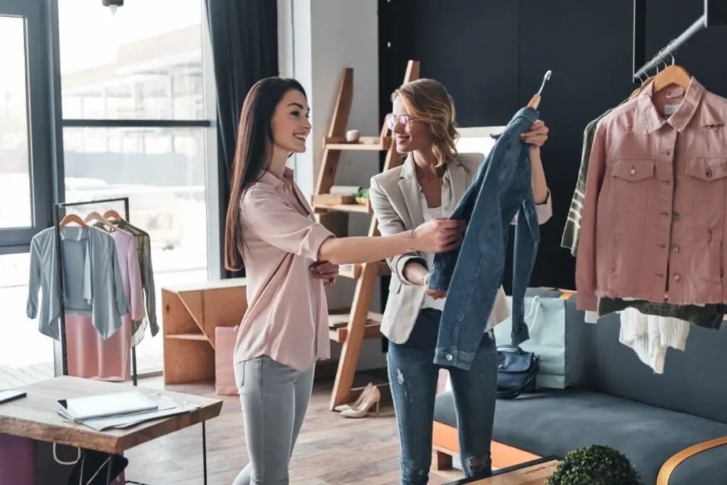Duas mulheres sorrindo em uma loja de roupas, segurando uma jaqueta jeans, ilustrando um atendimento de sucesso e a satisfação do cliente, o oposto de uma falha.