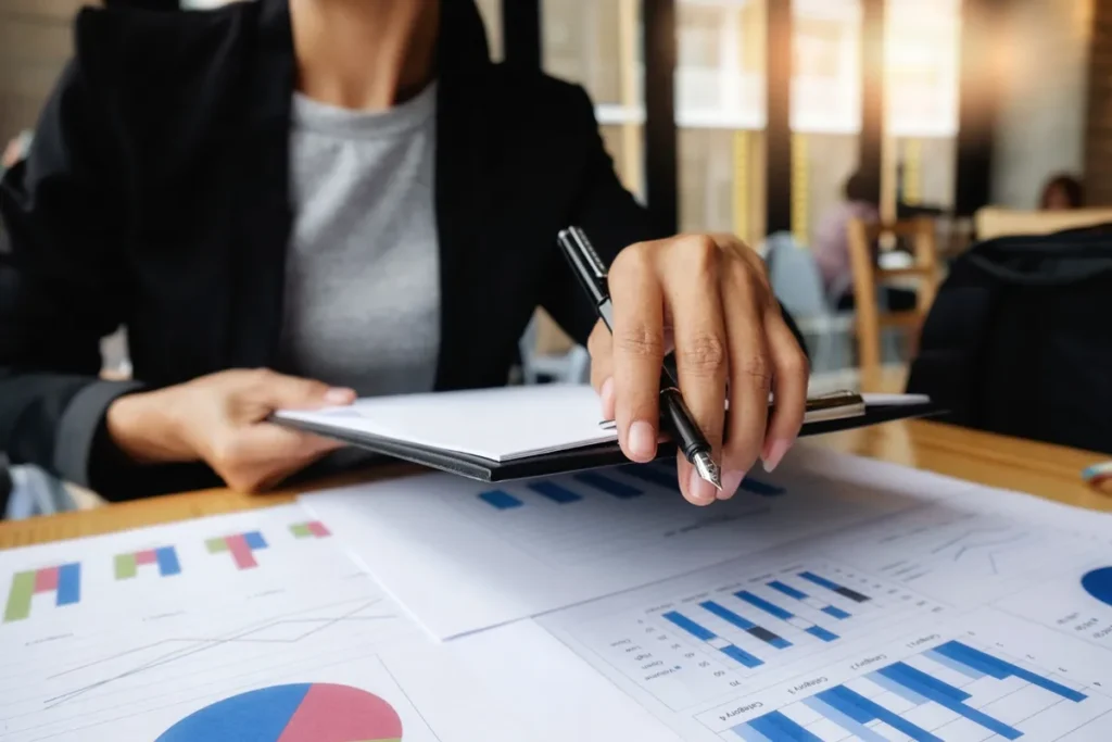 Mulher de negócios analisando gráficos e relatórios de faturamento e finanças, segurando uma prancheta e uma caneta, em uma mesa de escritório.