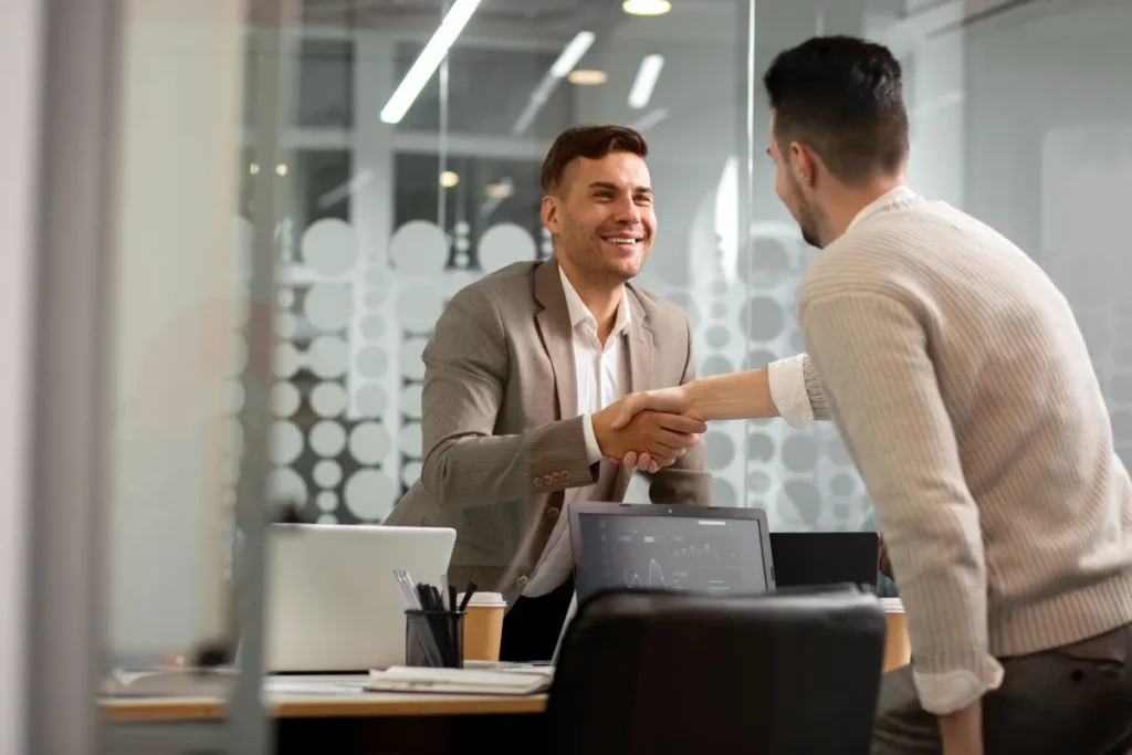 Dois homens apertando as mãos em um escritório moderno, com um laptop na mesa, simbolizando a finalização de um acordo.