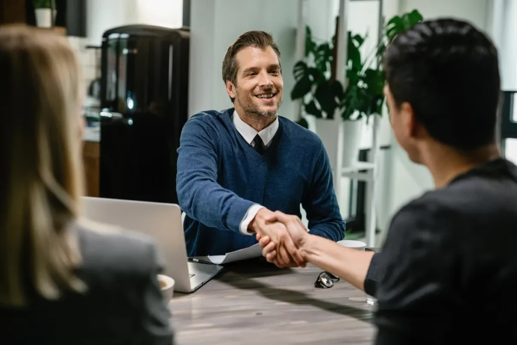 Homem de suéter azul sorrindo e apertando a mão de um cliente, em uma mesa com laptop e documentos.