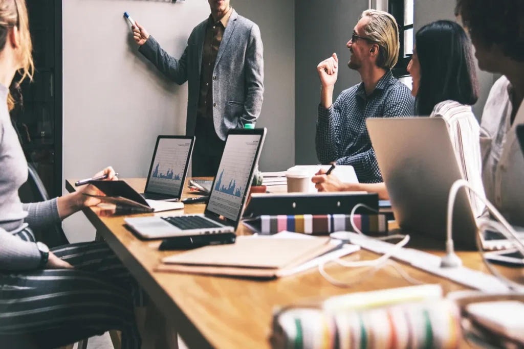 Uma equipe de negócios em uma reunião, com um homem apresentando dados de produtividade em um quadro branco, enquanto os colegas observam e usam laptops.