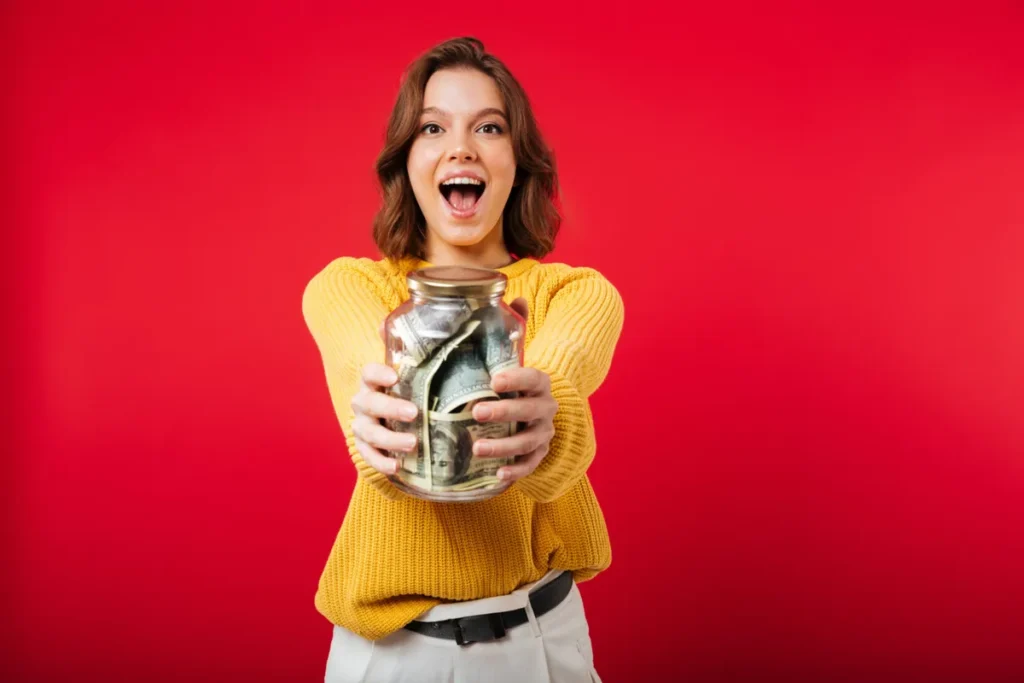 Mulher animada e sorrindo, segurando um grande pote de vidro cheio de dinheiro, simbolizando a conquista de renda extra e o sucesso financeiro