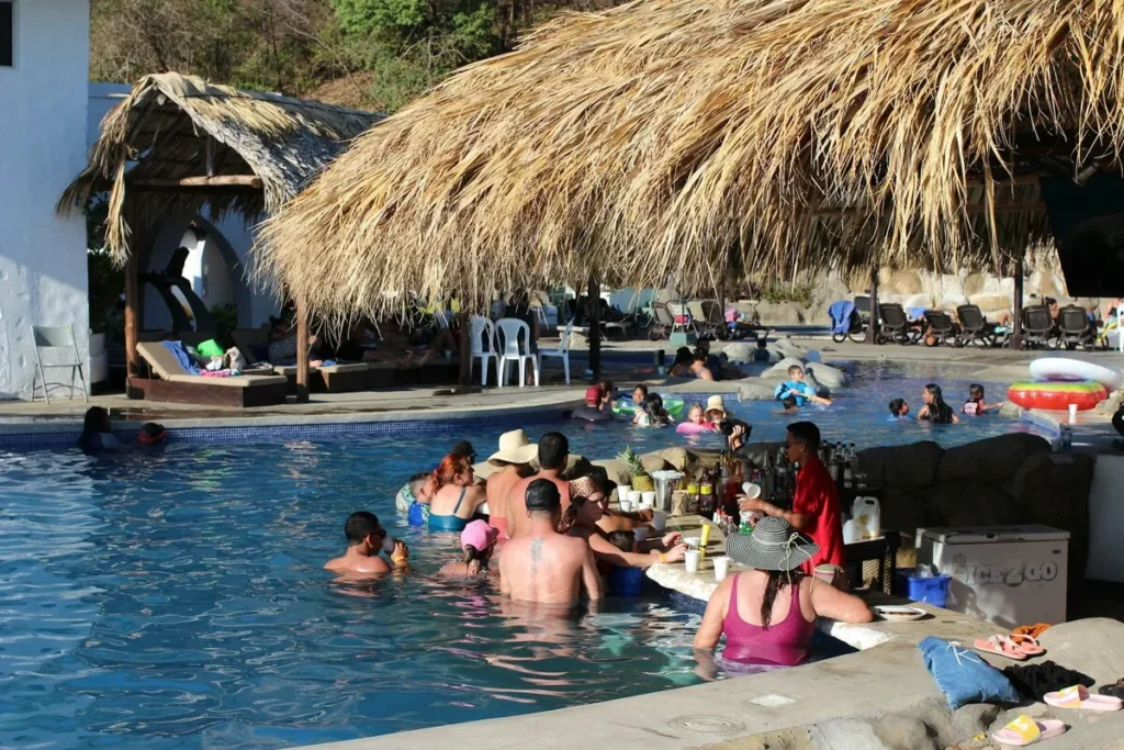 Bar molhado de piscina em resort all inclusive com teto de palha e hóspedes em trajes de banho interagindo e sendo servidos por um barman, sob sol forte.