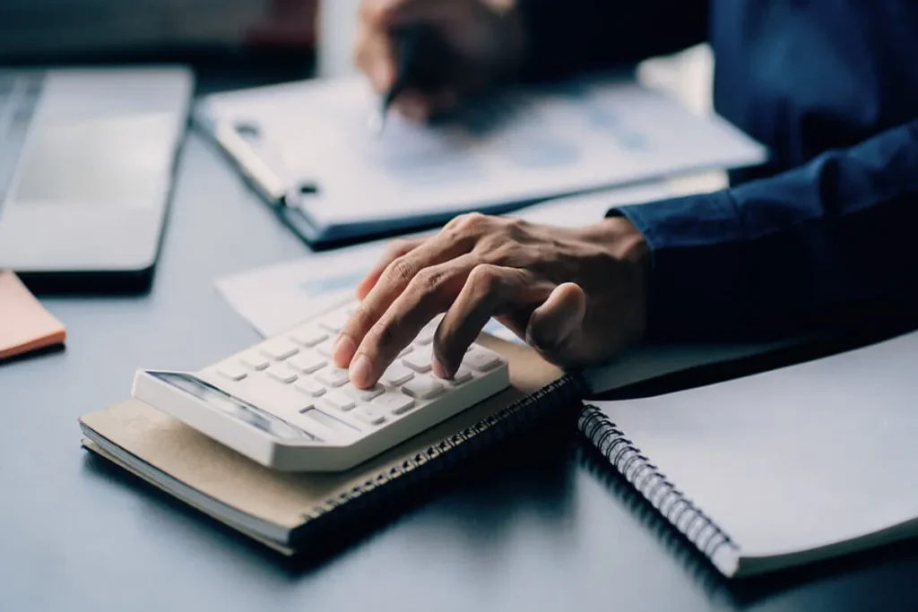 Uma pessoa usa uma calculadora e anotações para calcular a revenue e as finanças da sua empresa. A imagem representa o trabalho de análise financeira e planejamento para o crescimento da receita.