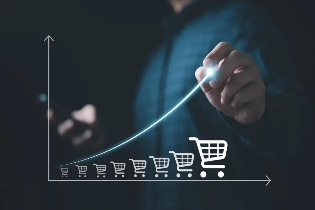 Uma mão toca uma tela, desenhando um gráfico de linha que cresce sobre uma série de carrinhos de compras que aumentam de tamanho. A imagem simboliza o aumento da revenue por meio do crescimento das vendas e da receita de um negócio.