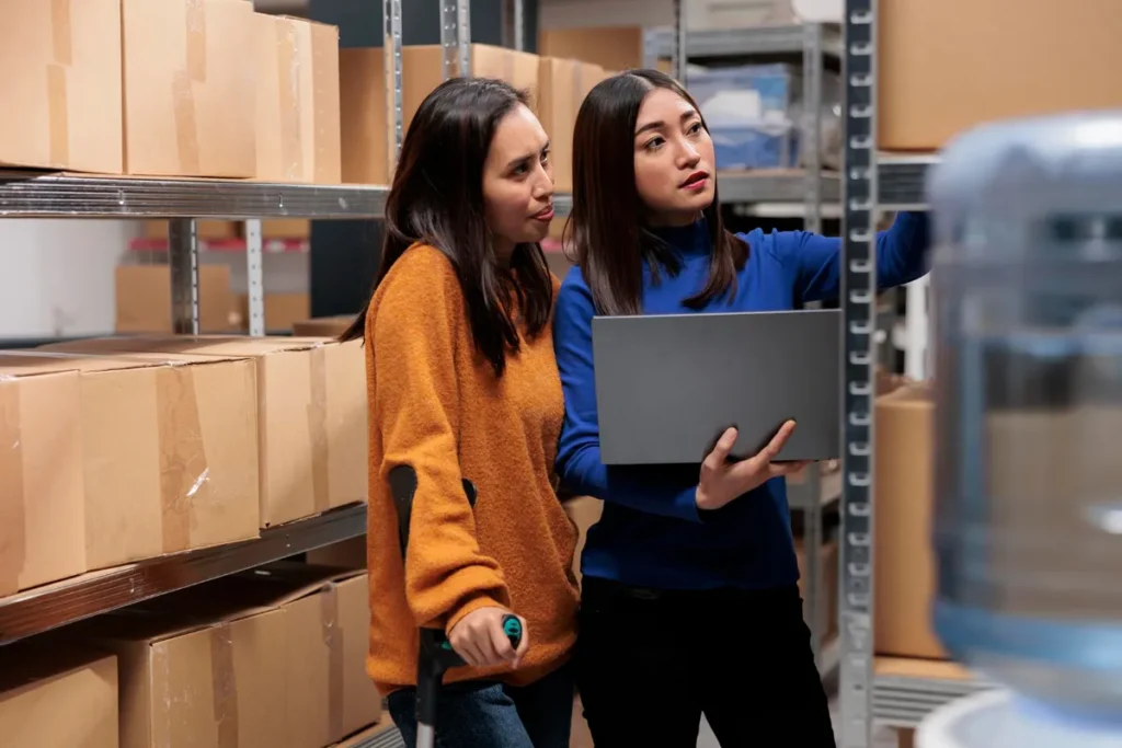 Duas mulheres em um armazém, uma delas com muletas, usando um laptop para checar o estoque e evitar a ruptura, em frente a prateleiras cheias de caixas.