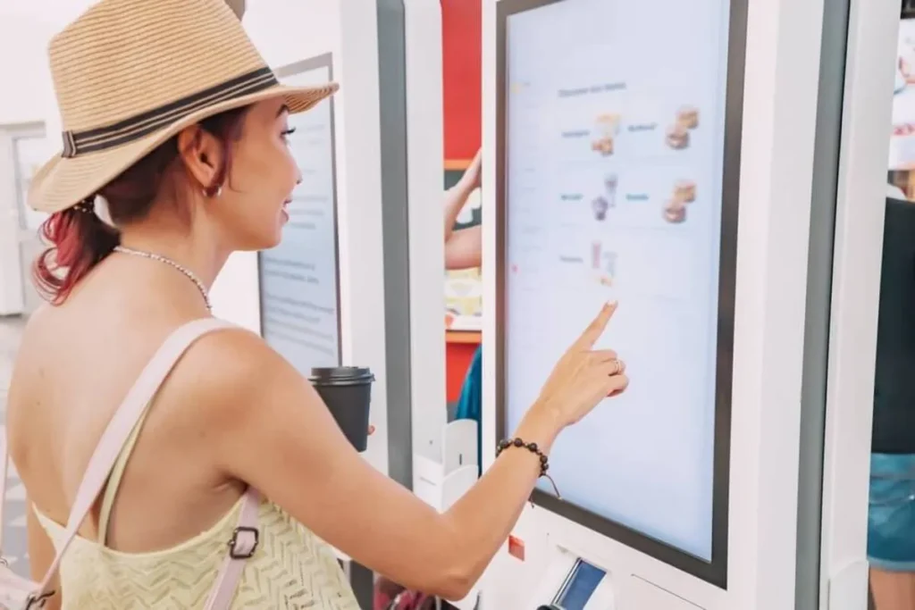 Mulher com chapéu de palha e copo de café, usando um totem de atendimento touchscreen para fazer um pedido, mostrando a praticidade do autoatendimento em restaurantes.