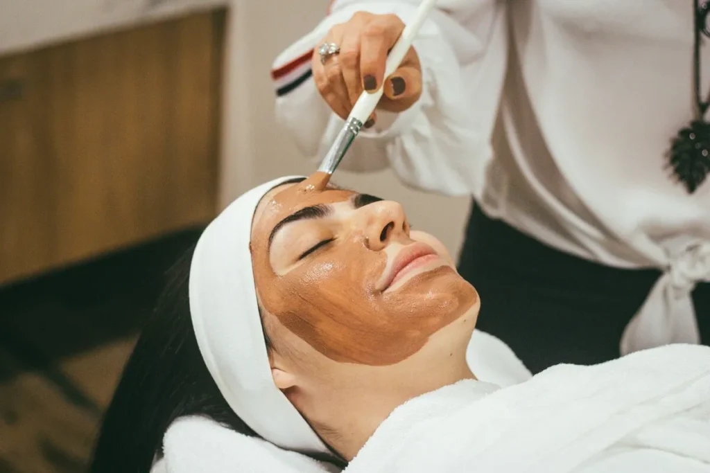 Uma mulher recebendo uma máscara facial com um pincel em um dos muitos SPAs (Serviços de Personalização e Atenção) que oferecem tratamentos estéticos. A imagem representa o cuidado com a pele e o relaxamento em ambientes especializados em beleza e bem-estar.