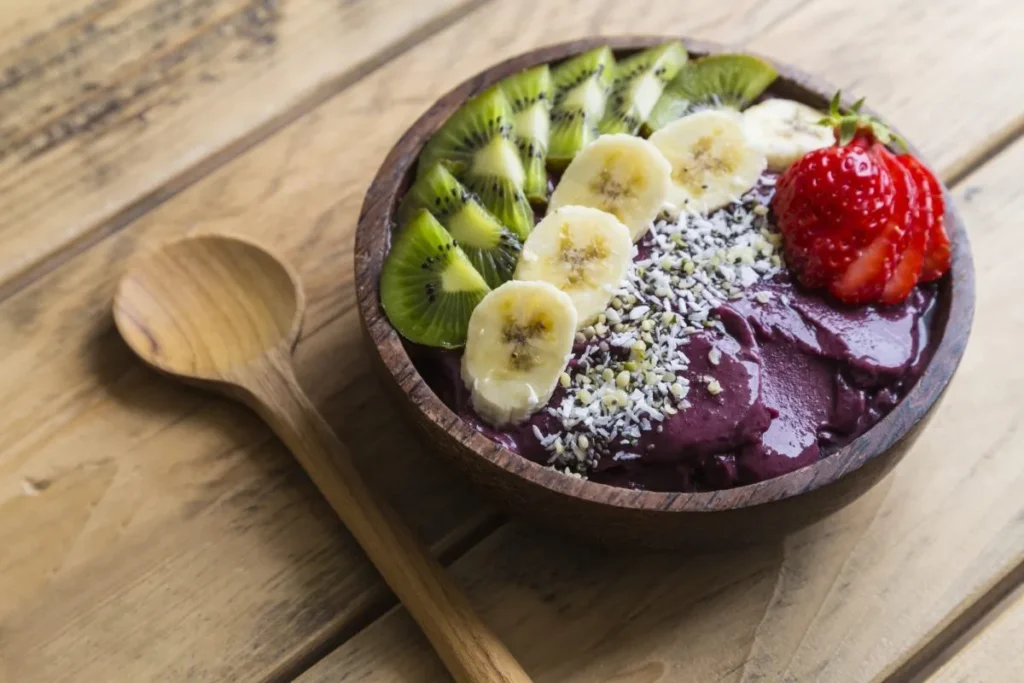 Tigela de açaí com fatias de kiwi e banana, morango cortado e sementes em uma mesa de madeira. Acompanhada por uma colher de madeira ao lado.