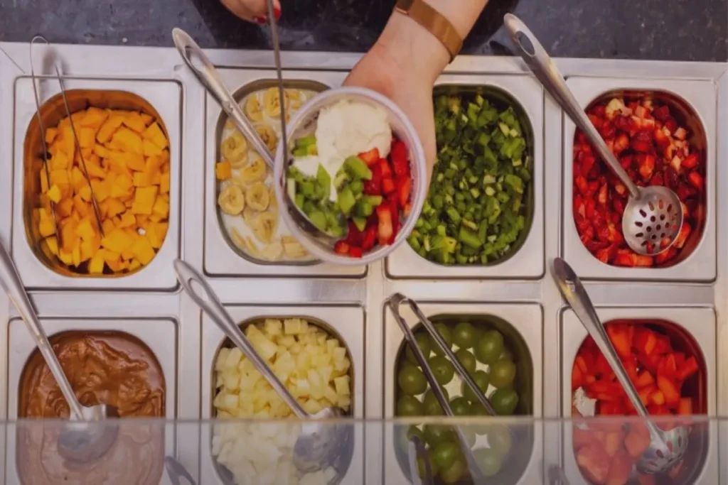 Açaíteria moderna com balcão de toppings variados, incluindo frutas como morango e banana, granola e Leite Condensado. Vendedor prepara bowl de açaí para cliente.