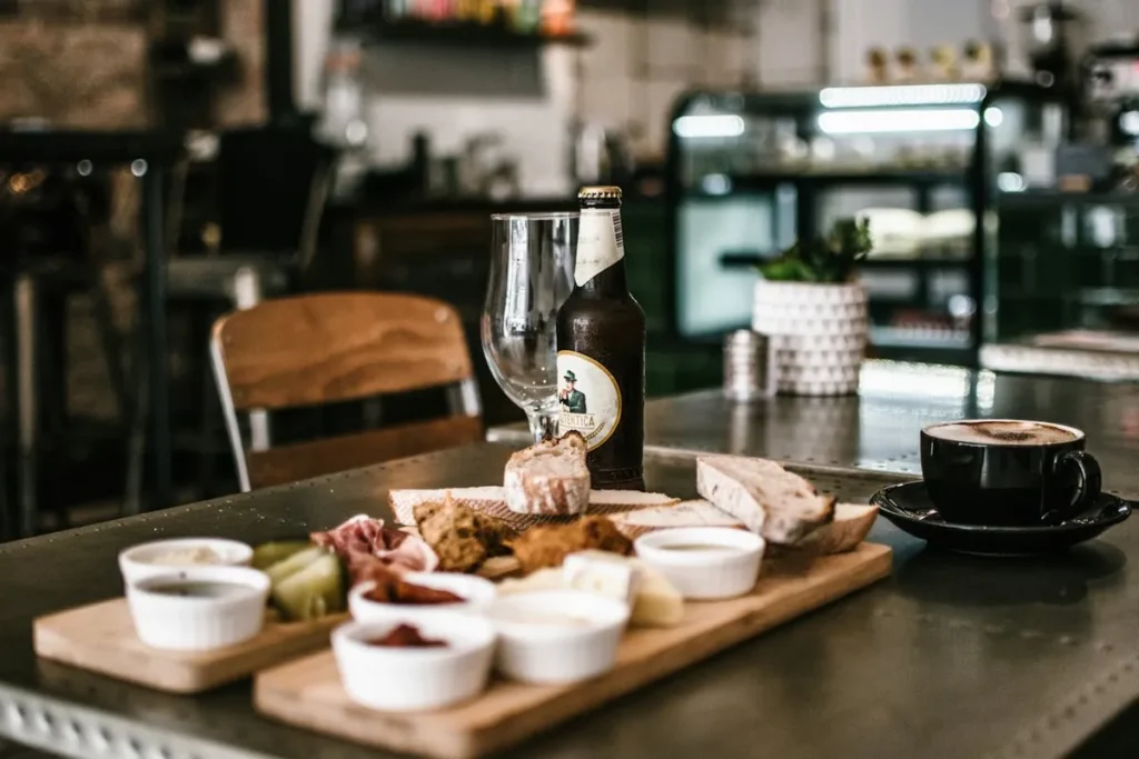 Mesa de bistrô com tábua de degustação de pães, queijos e frios, cerveja artesanal, taça de vidro e xícara de café, com ambiente interno moderno e decoração industrial no fundo.