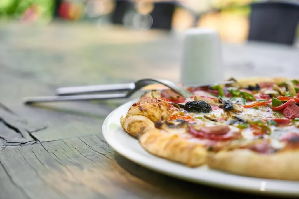 Close-up de fatia de pizza artesanal com vegetais, cogumelos e linguiça, servida em prato branco sobre mesa rústica de madeira em ambiente externo de bistrô.