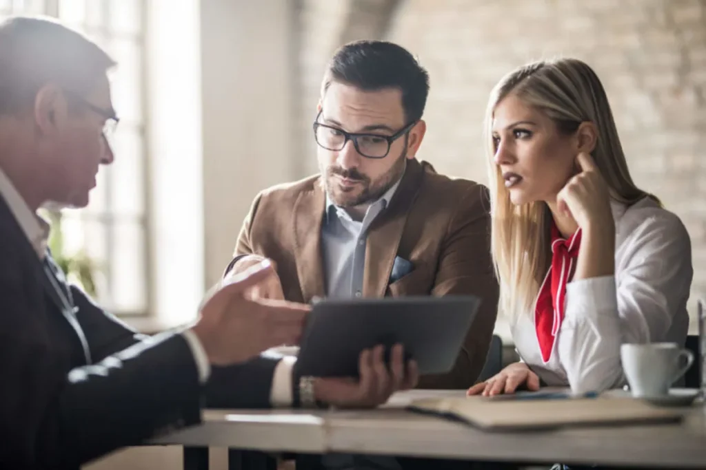 Um casal em uma reunião de negócios, conversando com um consultor que está mostrando informações em um tablet. A imagem representa o processo de um casal buscando uma corretora de seguro para entender e contratar um plano que atenda às suas necessidades.