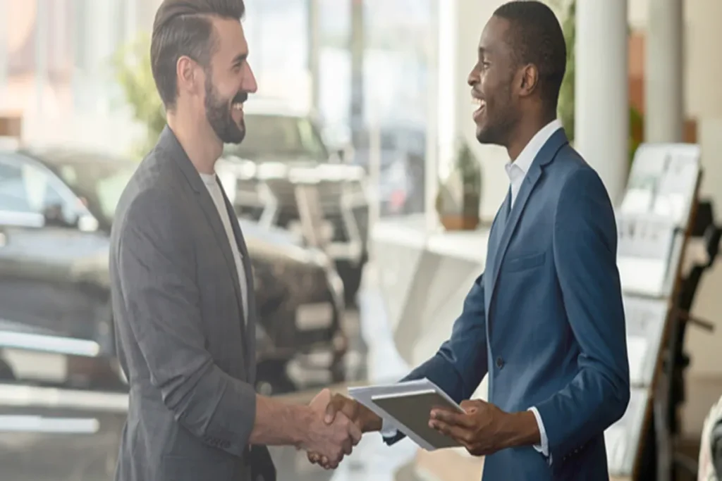 Dois homens de terno apertando as mãos em um showroom de carros, representando a negociação e a contratação de um seguro com a ajuda de uma corretora de seguro. A imagem ilustra a etapa de compra de um carro e a necessidade de proteção financeira.
